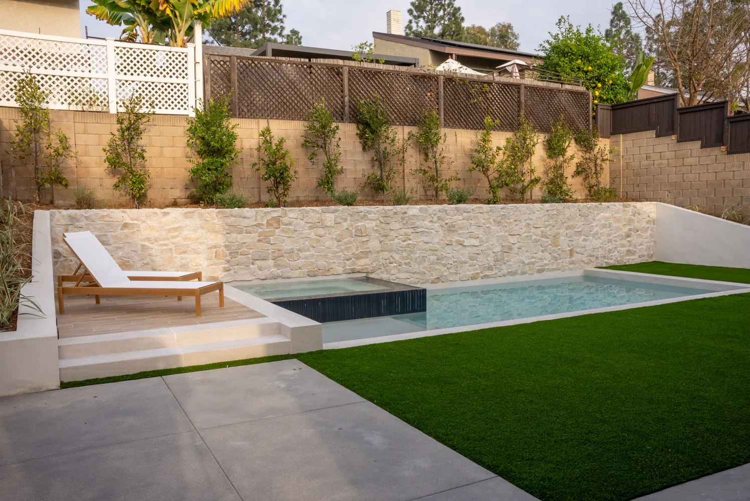 The pool and spa seen across a turf lawn from the patio, a lounger on the deck beyond.