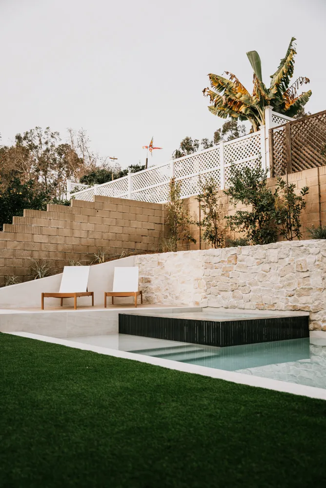 Two loungers above the dark spa, turf in the foreground and the retaining wall climbing behind.
