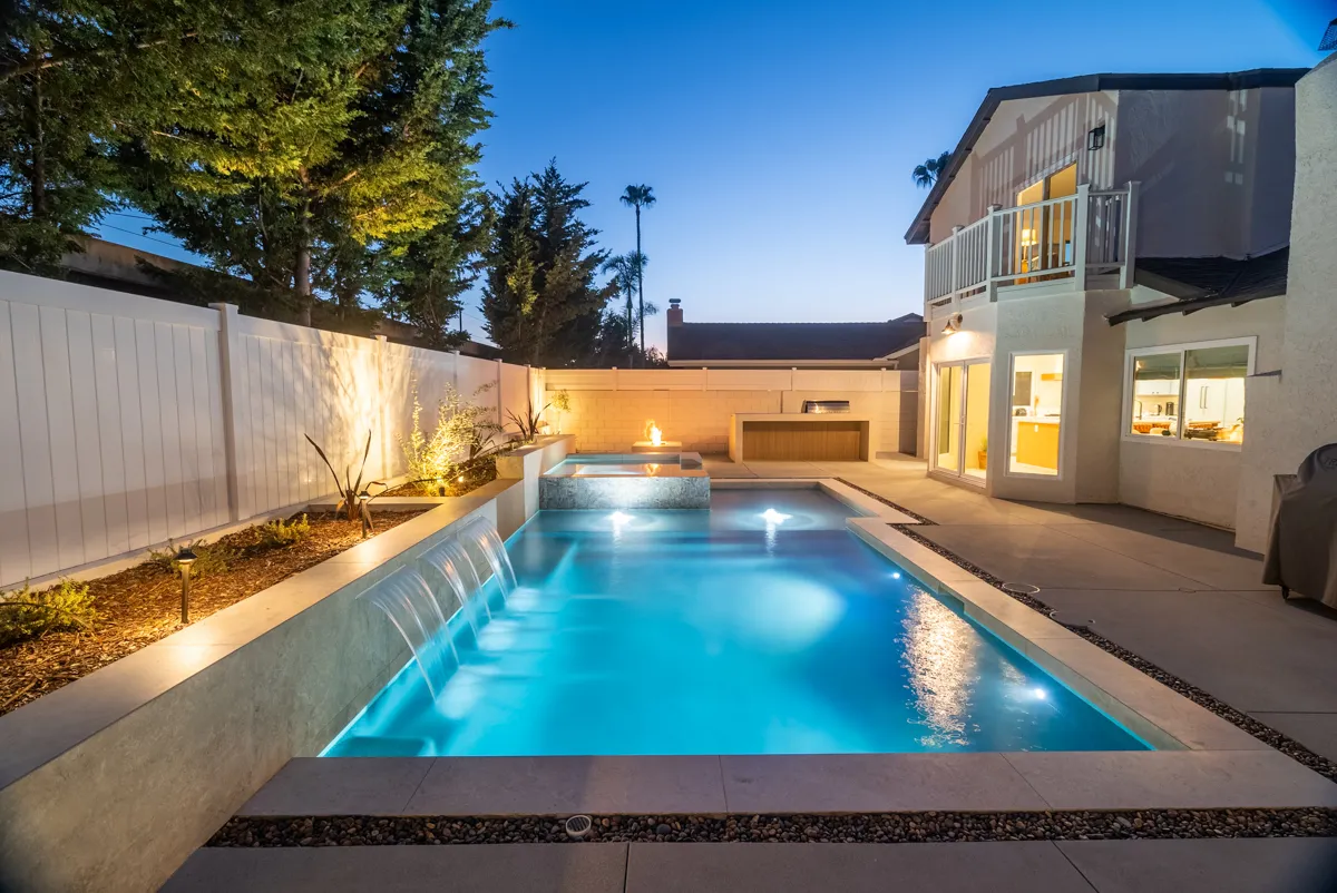 The pool lit at blue hour with a sheet of water falling from a raised wall, the house glowing behind it.