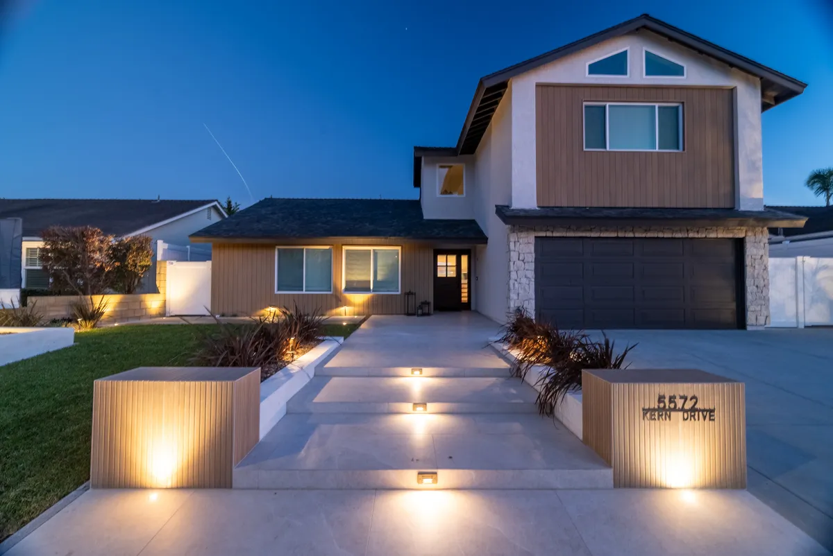 The front entry and driveway at blue hour, uplights in the paving and a lit address monument at the kerb.