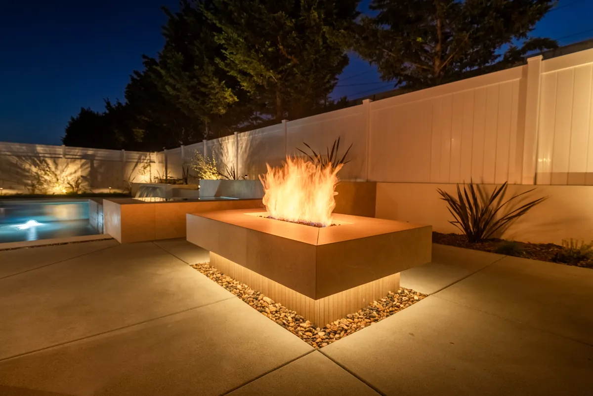 A square fire pit burning on the deck beside the pool, the wall behind it lit amber.