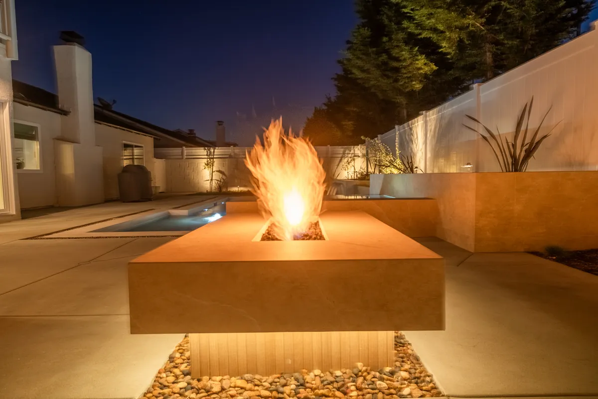 A tall flame rising from the fire pit against a deep blue sky, the pool and house behind.