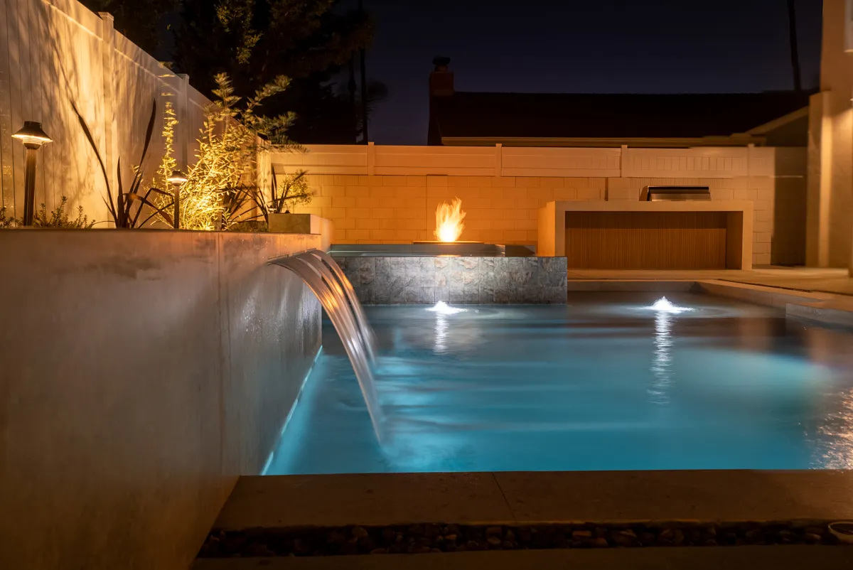 A wide sheet of water falling from the raised spa into the pool, with the fire pit burning beyond.