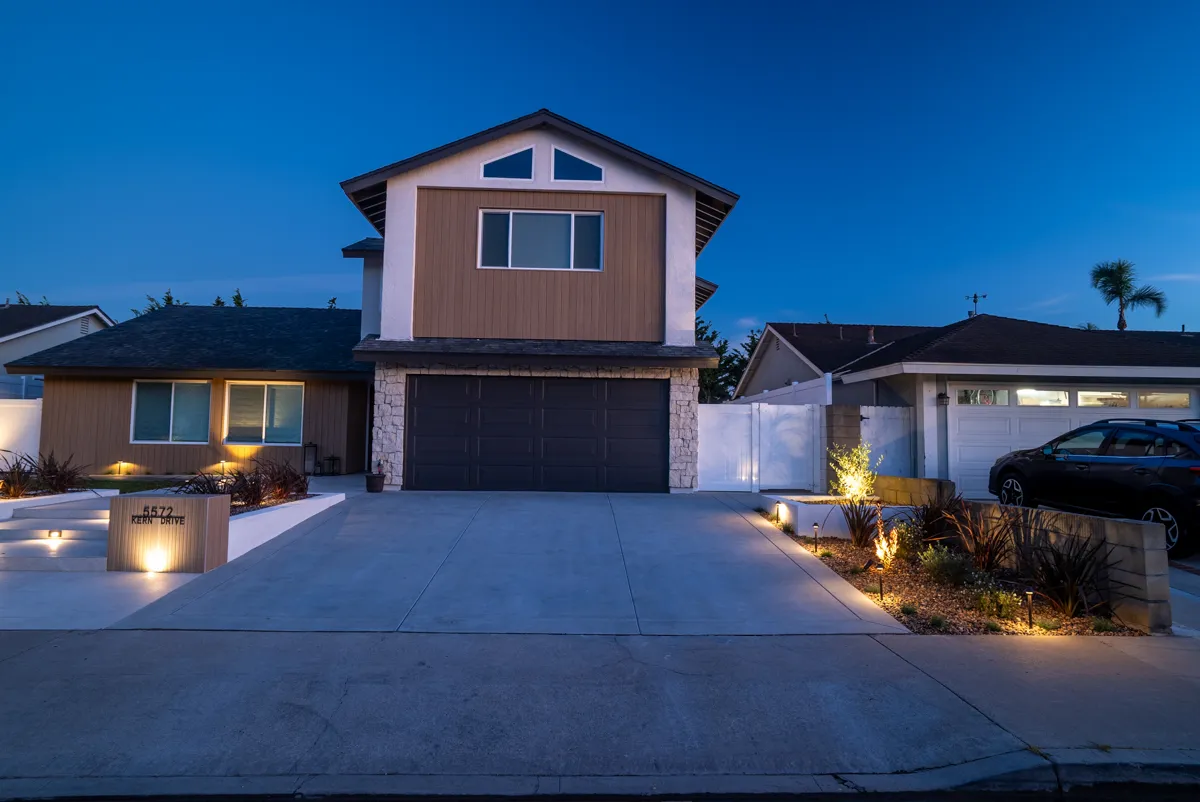 The house from the street at dusk, the driveway and garage washed with light and planting lit along the edge.