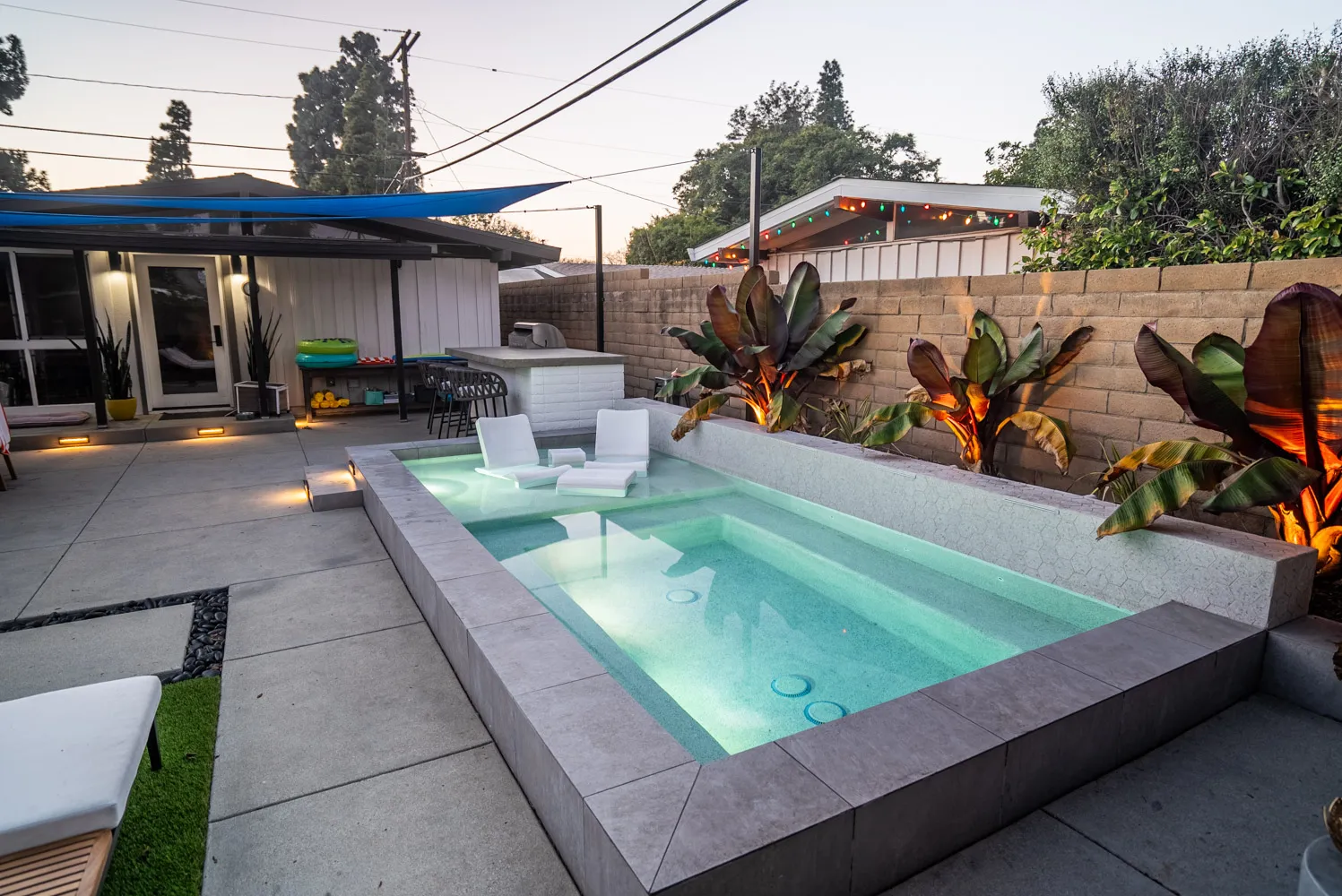 The pool from the deck at dusk, tropical planting along the wall and a low-slung house behind the shade sails.