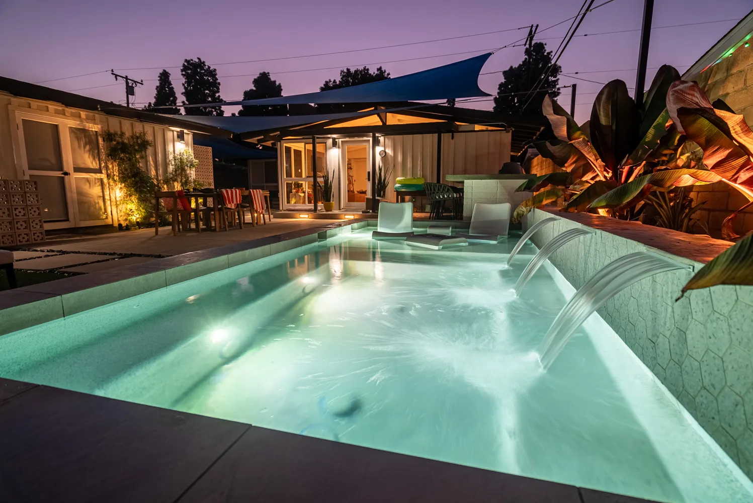 The pool lit turquoise under a violet sky, sheet falls running along the near wall and the house lit behind.