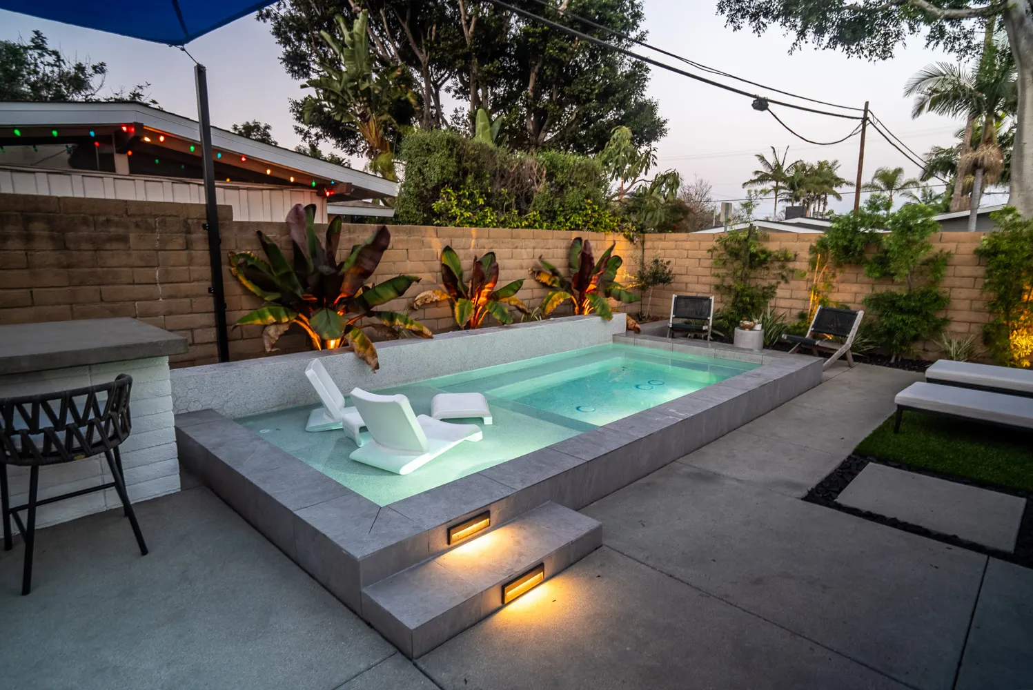 A raised pool with an in-water lounger on its shelf, blue shade sails overhead and a block wall planted with red-leaved bananas behind.