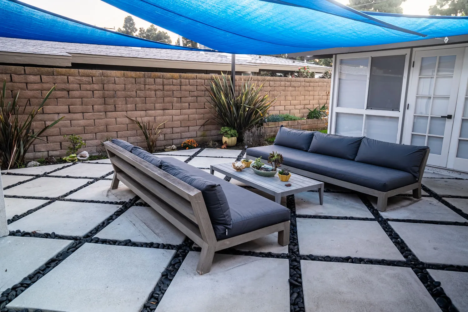 Two low sofas facing across a timber table on wide paving set into gravel, under the blue shade sail.