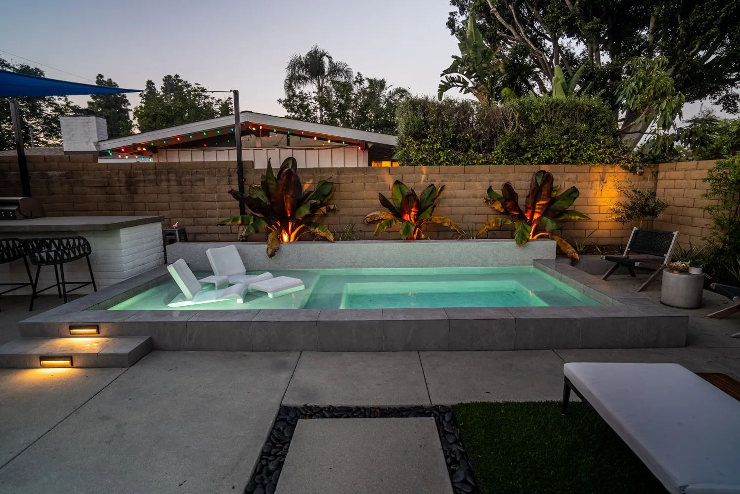 The whole yard at dusk, with a raised pool, loungers, planted wall and the bar counter at the near end.