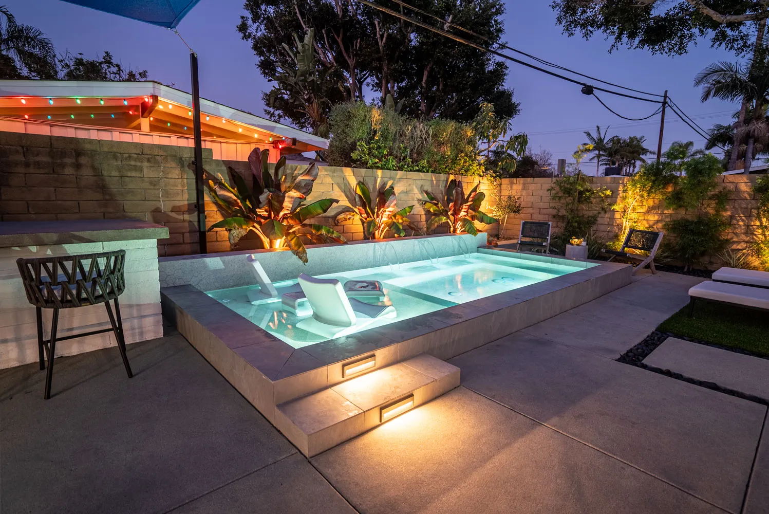 The lit pool at blue hour with an in-water lounger, the planted wall glowing behind and string lights along the eaves.