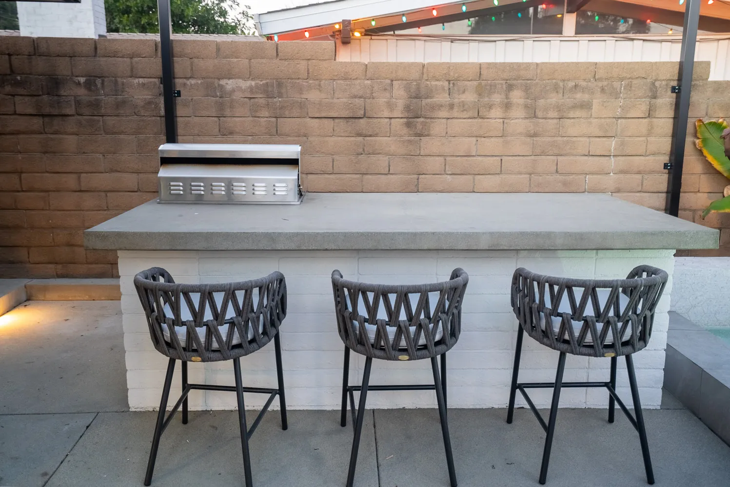 An outdoor bar counter with a built-in grill and three woven stools set against a block wall.