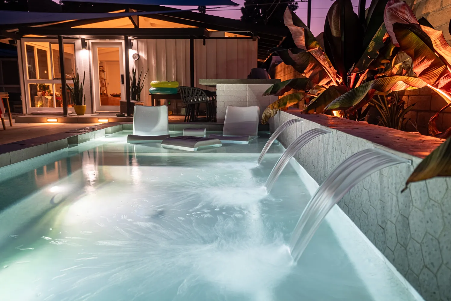 Sheet waterfalls falling into the pool after dark, the lit house standing open at the far end.