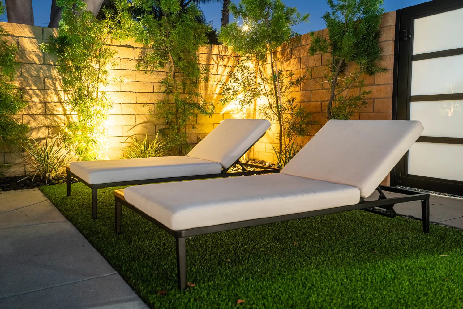 Loungers on the turf after dark, the wall behind washed with warm light through the planting.