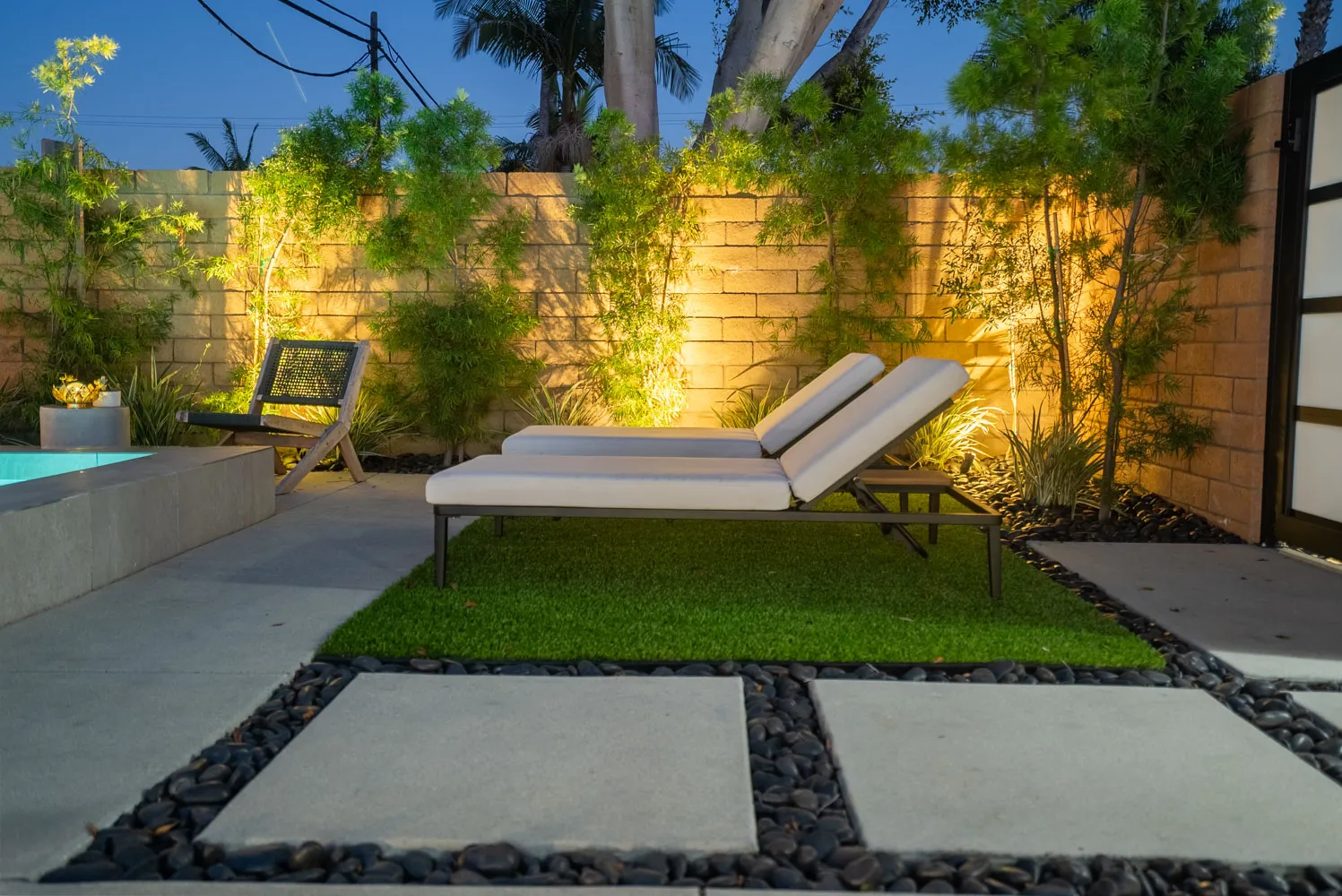 Two loungers on turf beside pale paving set into gravel, planting uplit against the block wall.