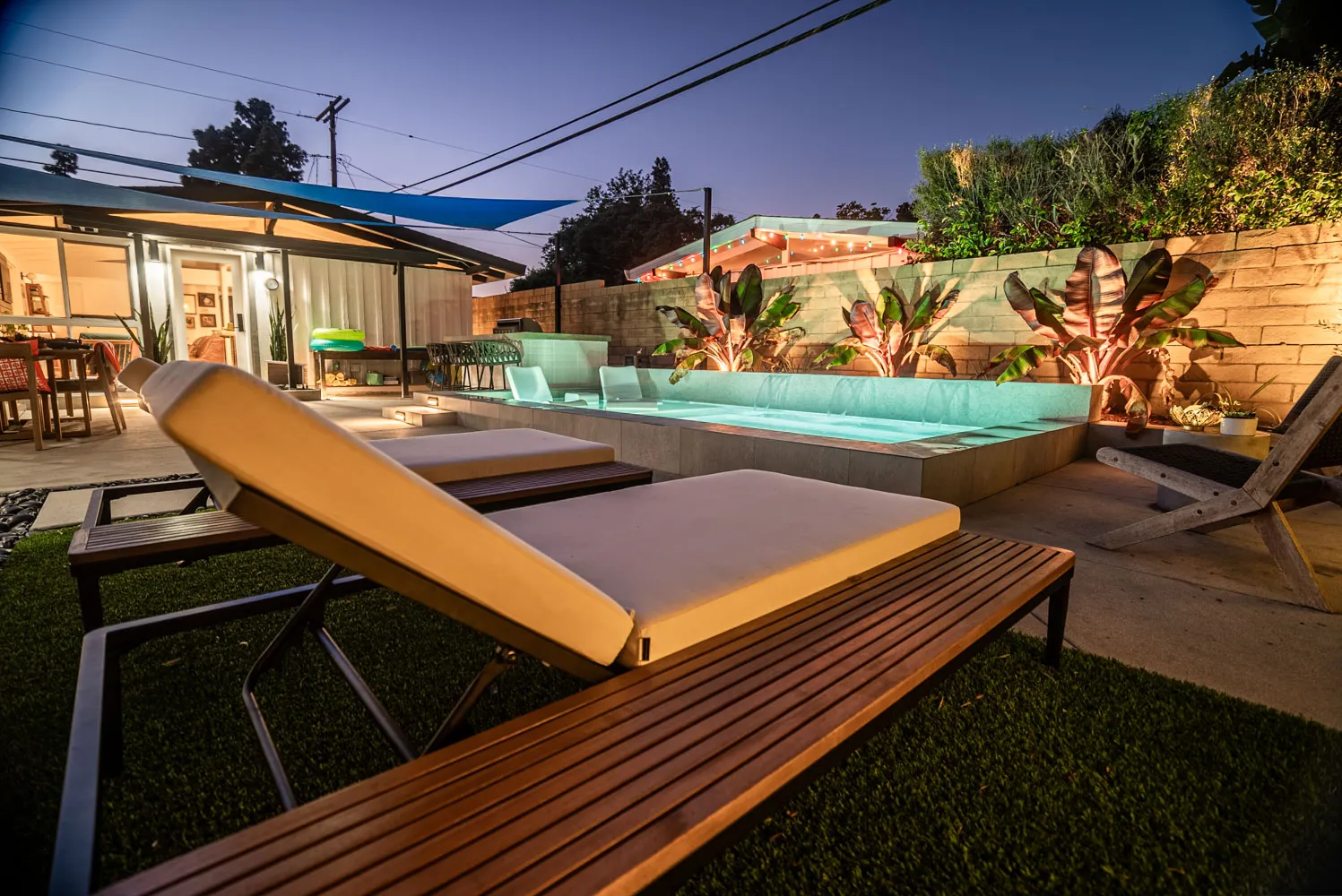 A timber lounger on the turf at dusk, the lit pool and planted wall running away behind it.