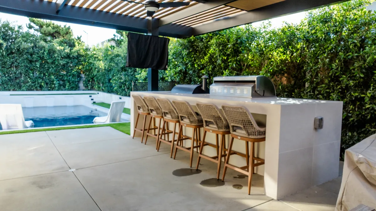 A bar counter with woven stools under a slatted pergola, the pool and clipped hedge running away behind it.