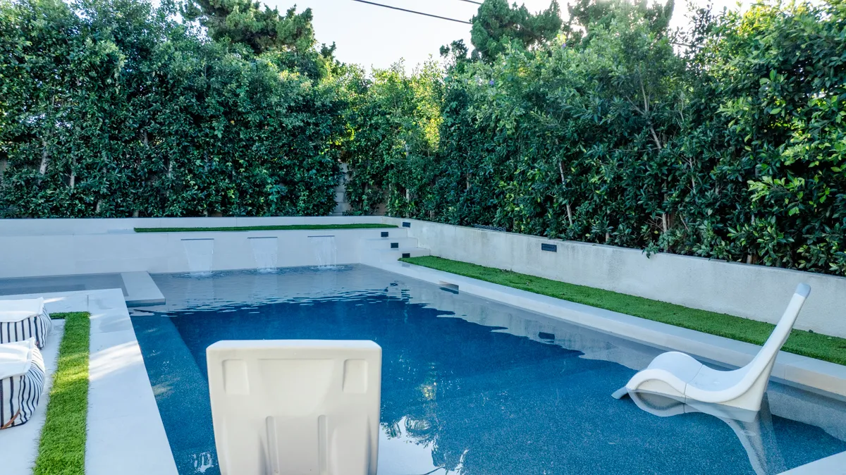 A white lounger standing in the shallow end, striped cushions on the deck and hedge filling the frame behind.