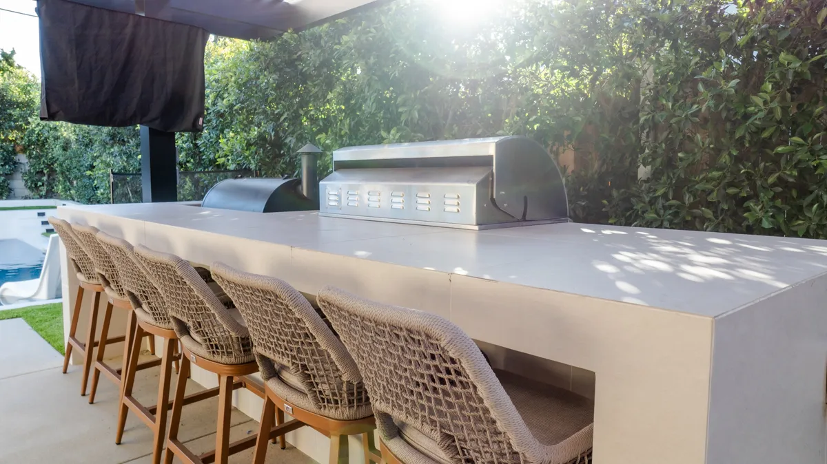 The outdoor kitchen counter with a built-in grill and warming drawers, woven stools drawn up along it.