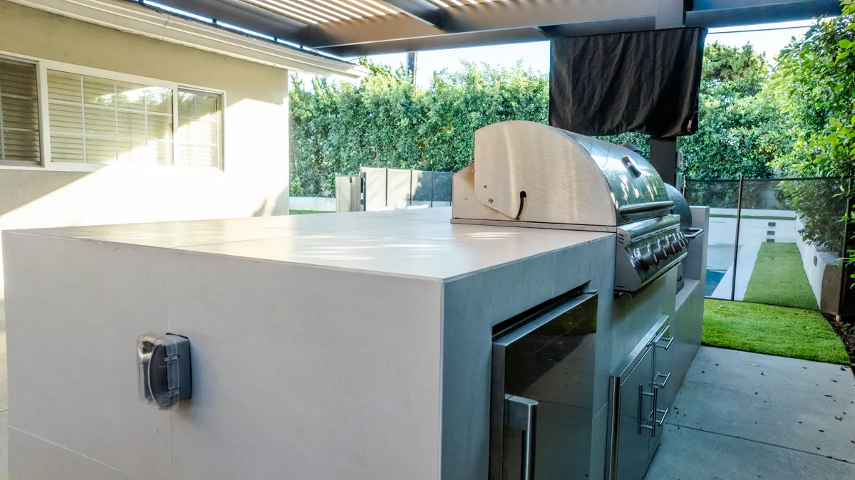 Close on the grill set into the poured counter, the house wall and glass fence behind.