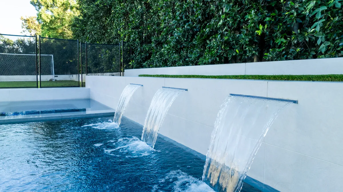 Three sheet waterfalls falling from a raised wall into the deep end, glass fencing and hedge above.