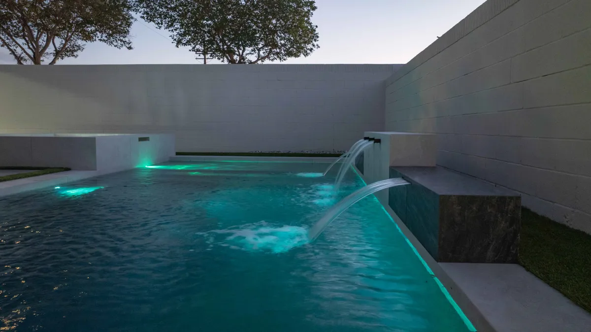 The pool lit green at dusk, a single sheet of water falling from the raised wall and trees standing over the boundary.