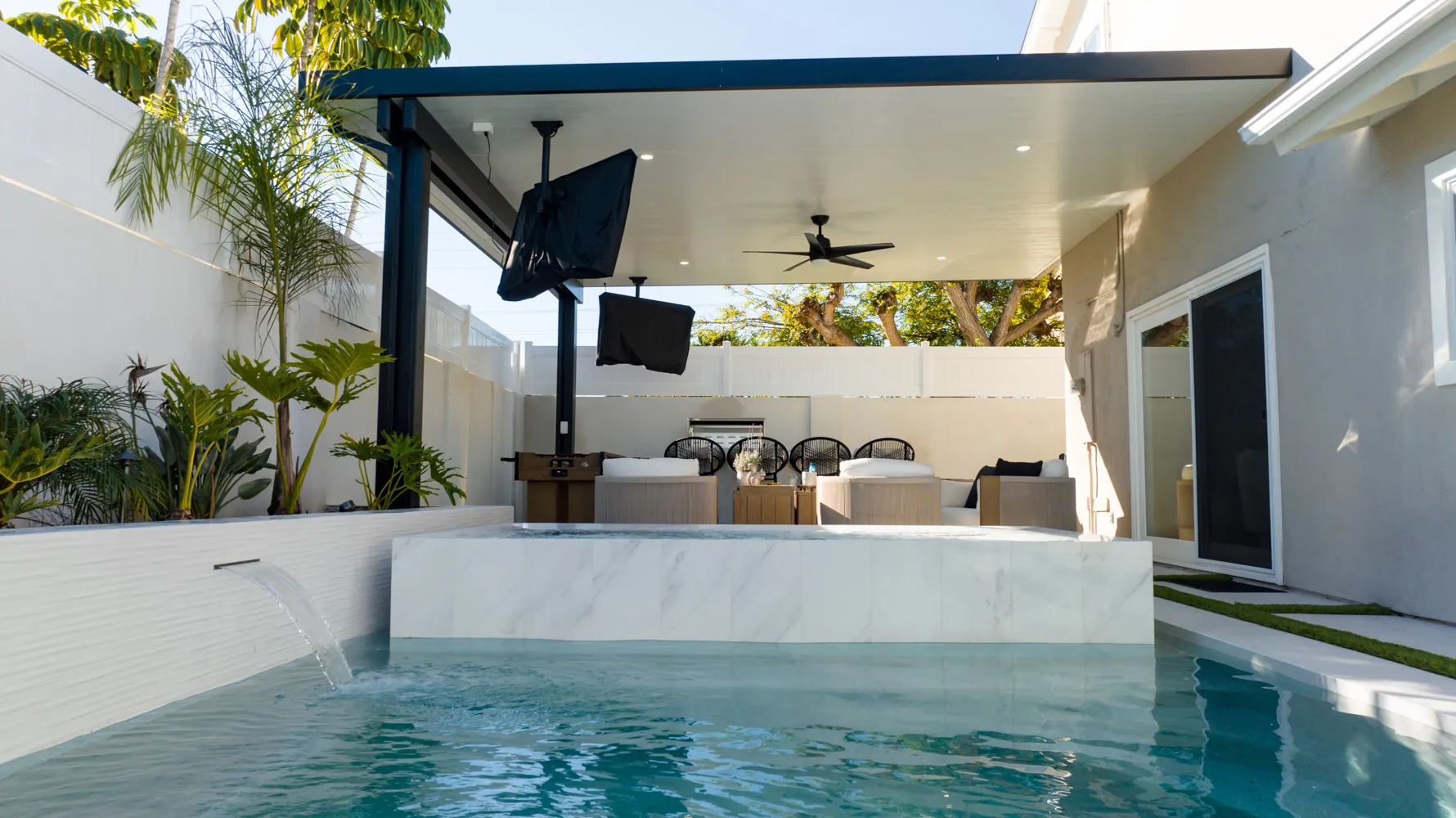 A scupper spilling into the pool from a marble-faced wall, the covered seating area and dining table beyond.