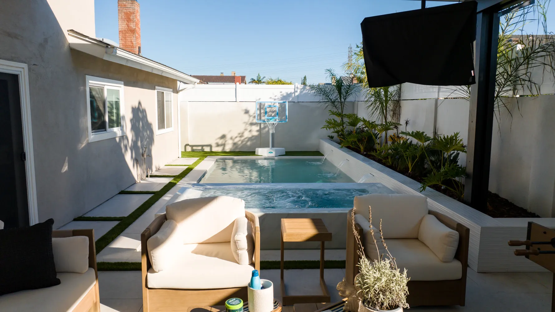 Cushioned chairs on the deck beside the pool, the house on one side and a television mounted under the patio cover.