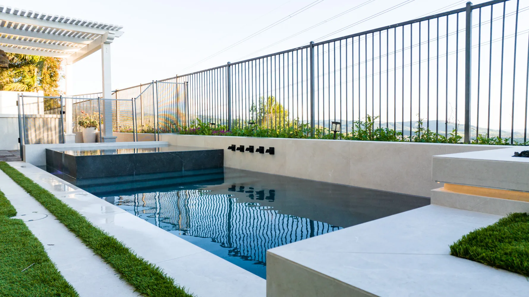 A long, narrow pool running the length of a ridge-top yard, a pergola at the near end and an open metal fence along the far side.