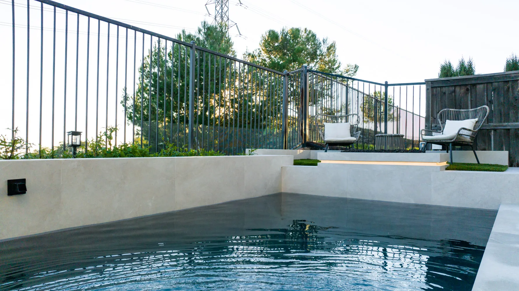 The pool from the far corner, a low rendered wall on one side and mature trees standing past the open fence.