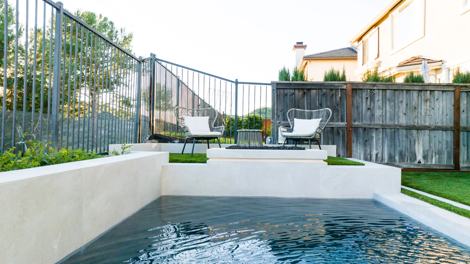 The shallow end with two woven chairs set on the turf, a timber fence and neighbouring rooflines behind.