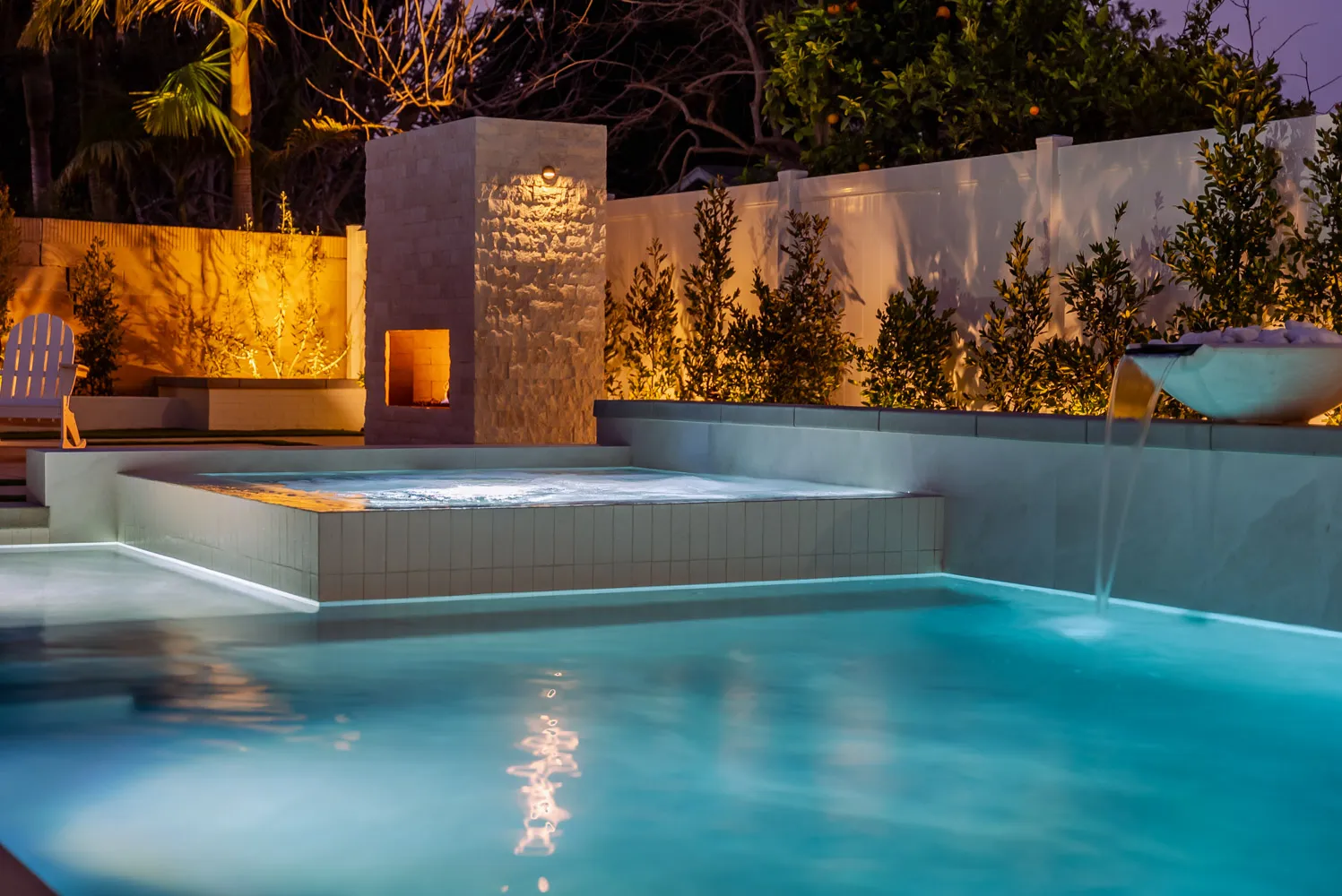 A raised spa spilling into the pool at dusk, with a stone-clad fireplace tower lit behind it and the far wall washed amber.