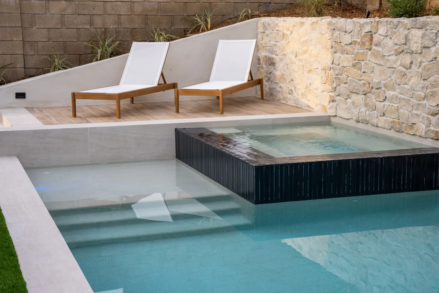 A pool set into turf below a stacked-stone retaining wall, with a dark-tiled raised spa spilling into the shallow end and two teak loungers above it.
