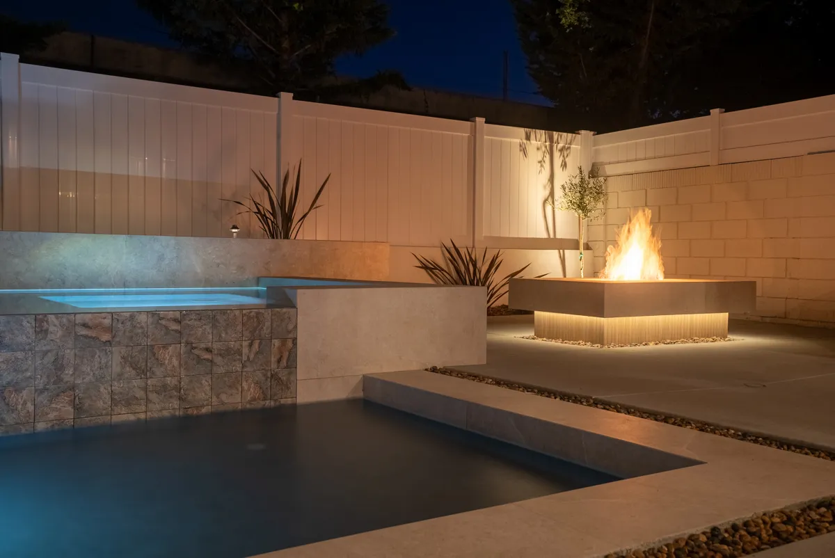 A lit pool at blue hour with a square fire feature burning at the far end and a stucco wall washed amber behind it.
