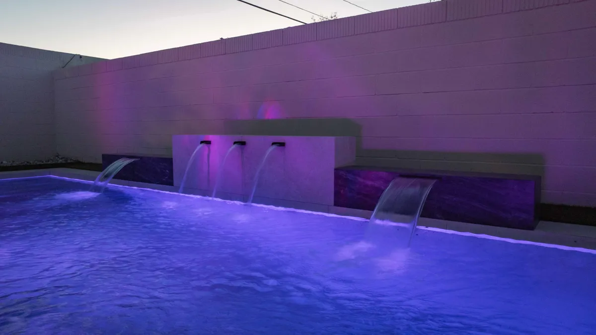 A pool lit violet after dark, the colour washing up a plain rear wall behind two sheet waterfalls.