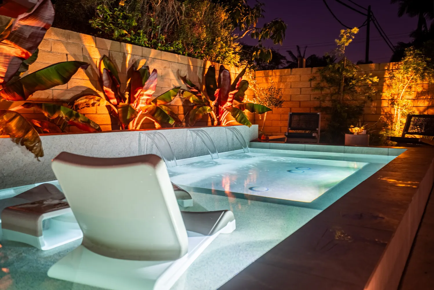 A lit pool at dusk under a violet sky, edged with tropical planting and a cushioned lounger set close to the water.