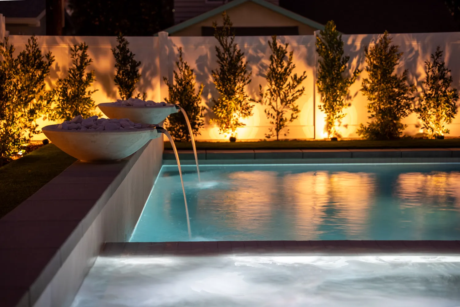 A rectangular pool at dusk with a stone bowl spilling water into the shallow end, behind it a row of slim trees uplit against a white wall.
