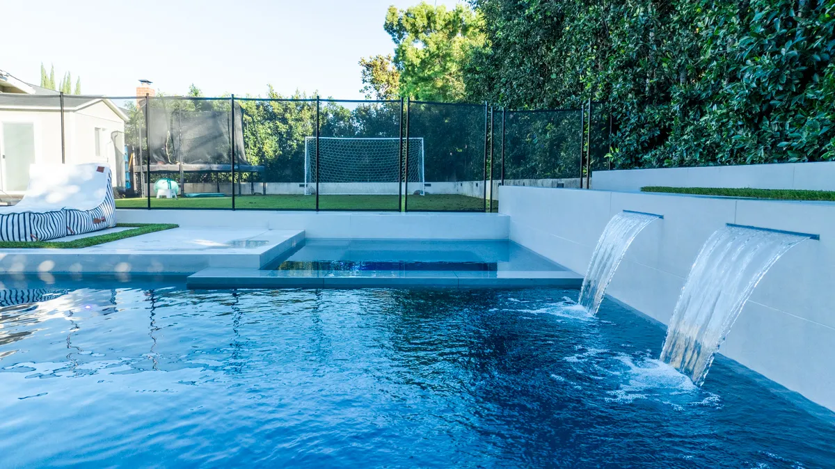 A deep blue pool enclosed by a tall clipped hedge, with two sheet waterfalls falling from a raised wall into the deep end.