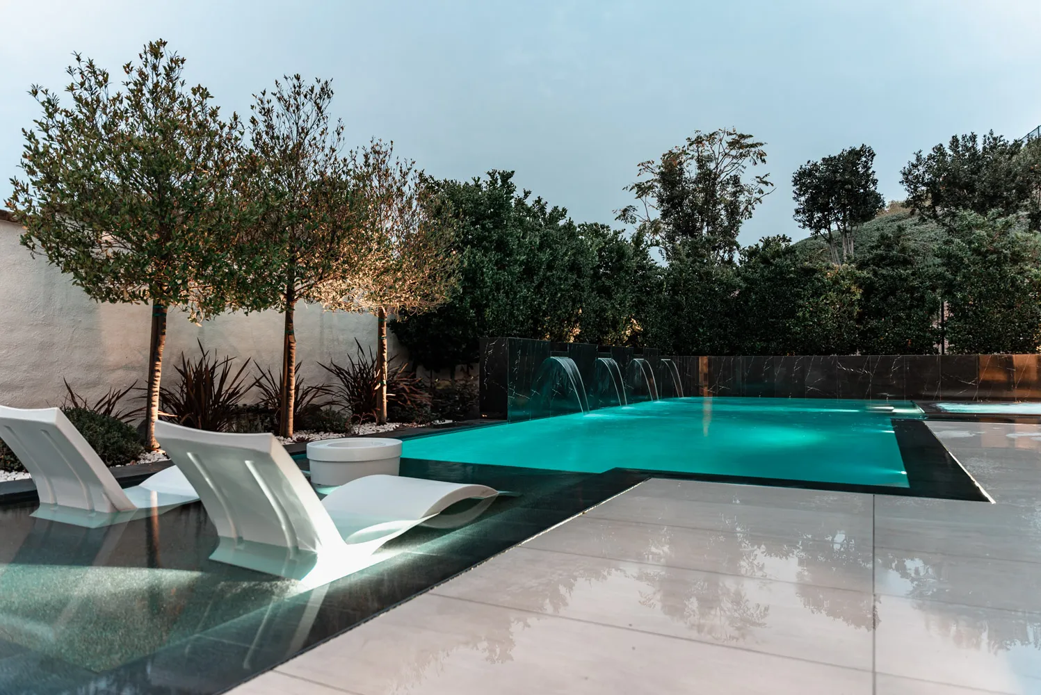 A vanishing-edge pool at dusk, its far lip level with a treeline, with white loungers and young olive trees on the deck beside it.