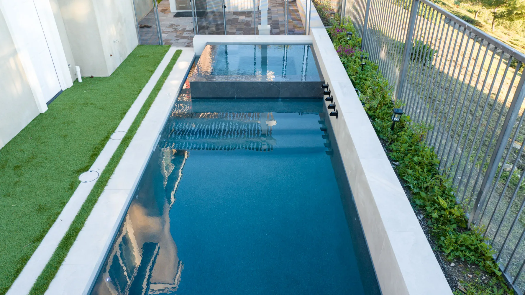 A long, narrow pool running the length of a ridge-top yard, with a strip of turf on one side and an open metal fence on the other looking out over trees.
