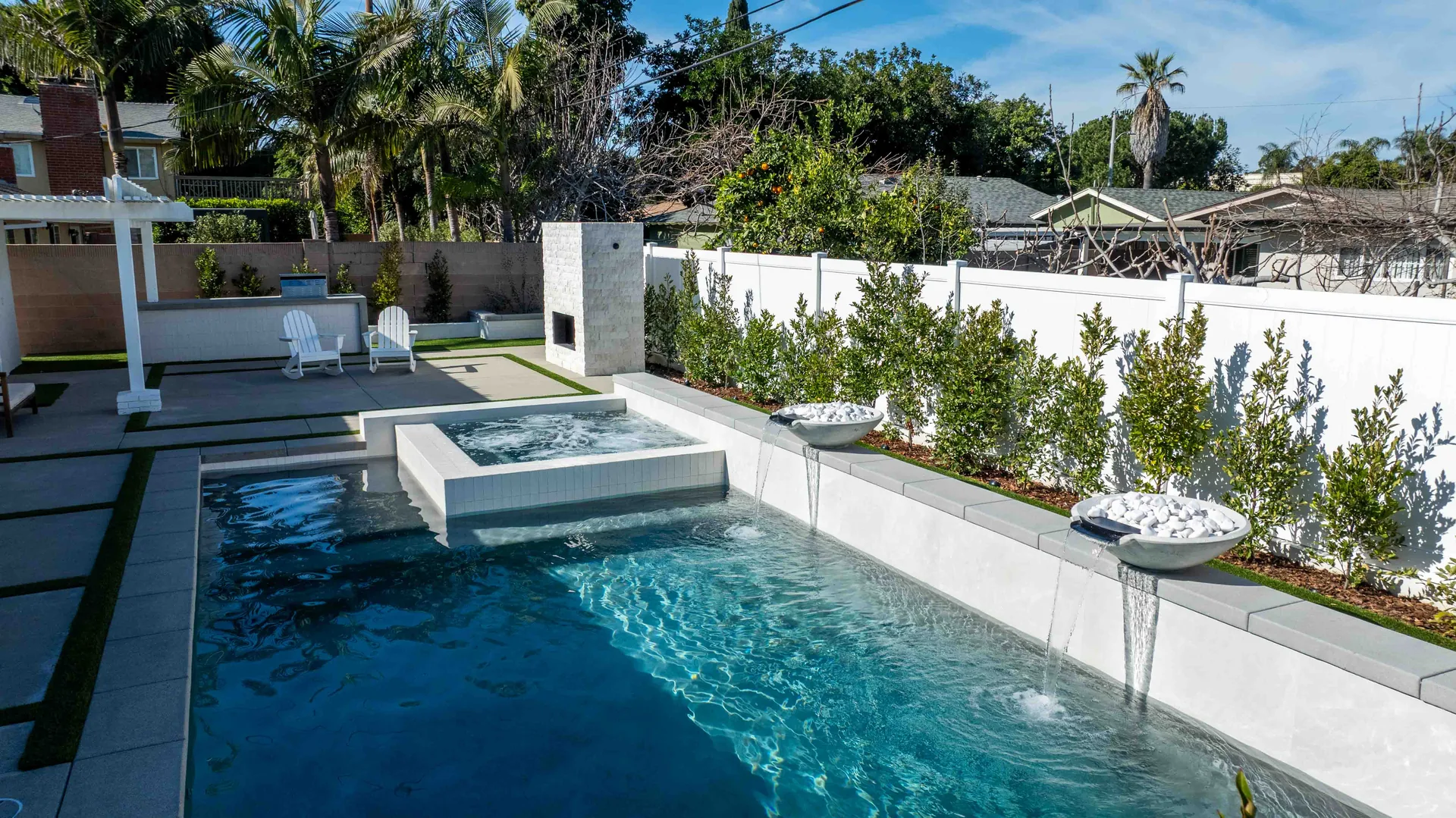 The full length of the pool in daylight, a raised spa at the near end and a row of young trees running the length of the white boundary wall.