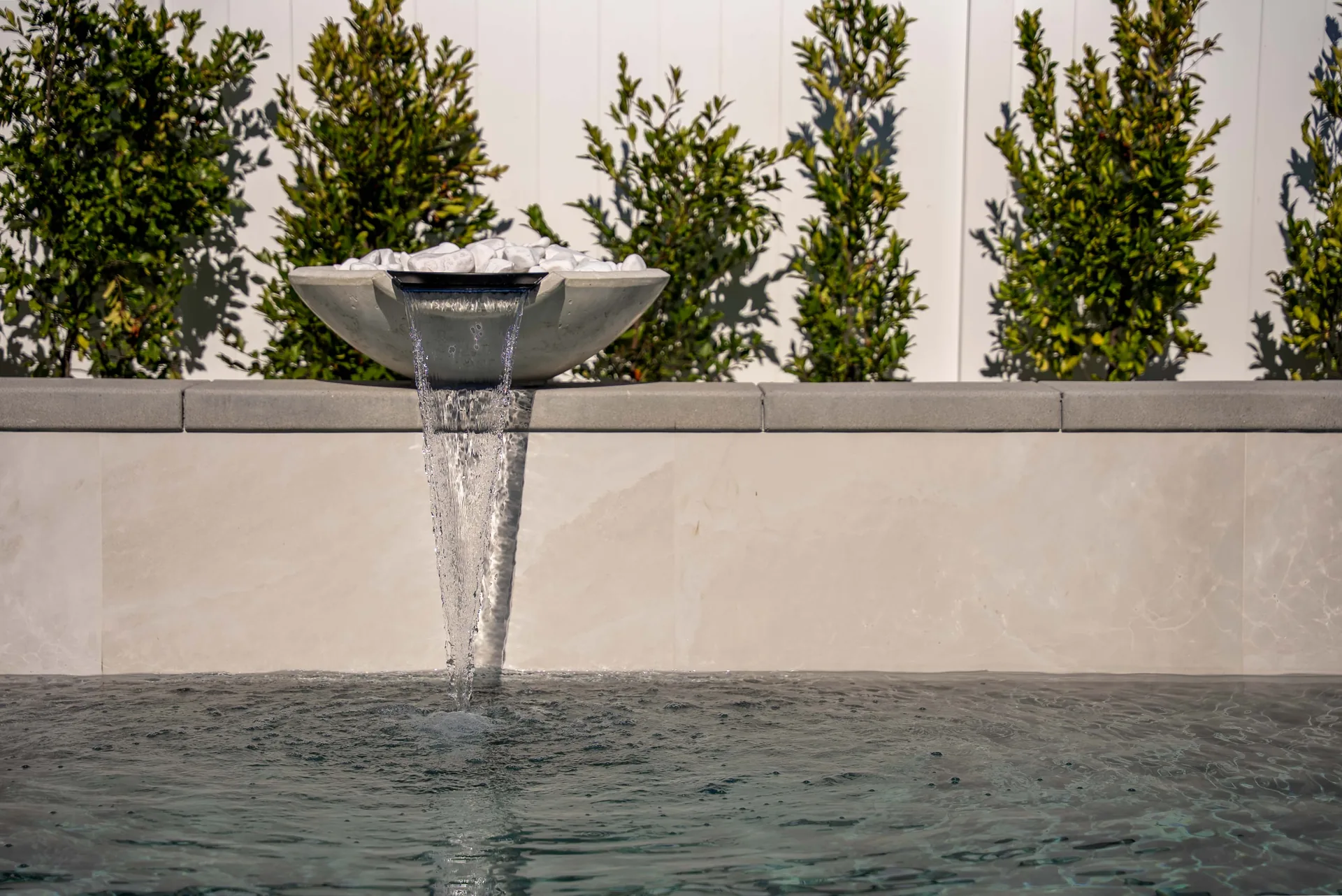 A wide sheet of water falling from a shallow bowl into the pool, against a wall planted with young trees.