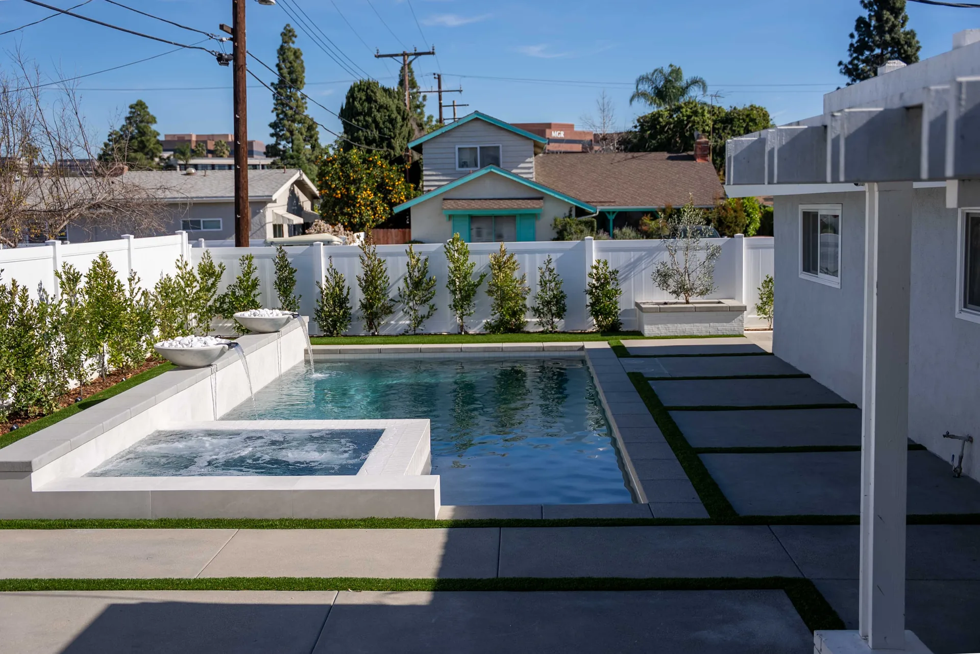 The pool and spa in flat daylight, paver strips crossing the deck and neighbouring rooflines above the wall.
