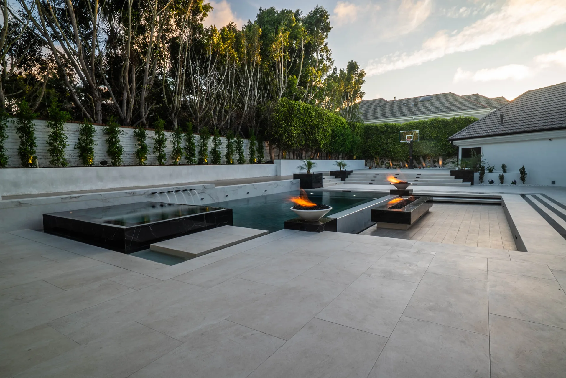 A dark-tiled pool set flush into pale stone paving, black fire bowls burning along its far edge and a clipped green wall behind.
