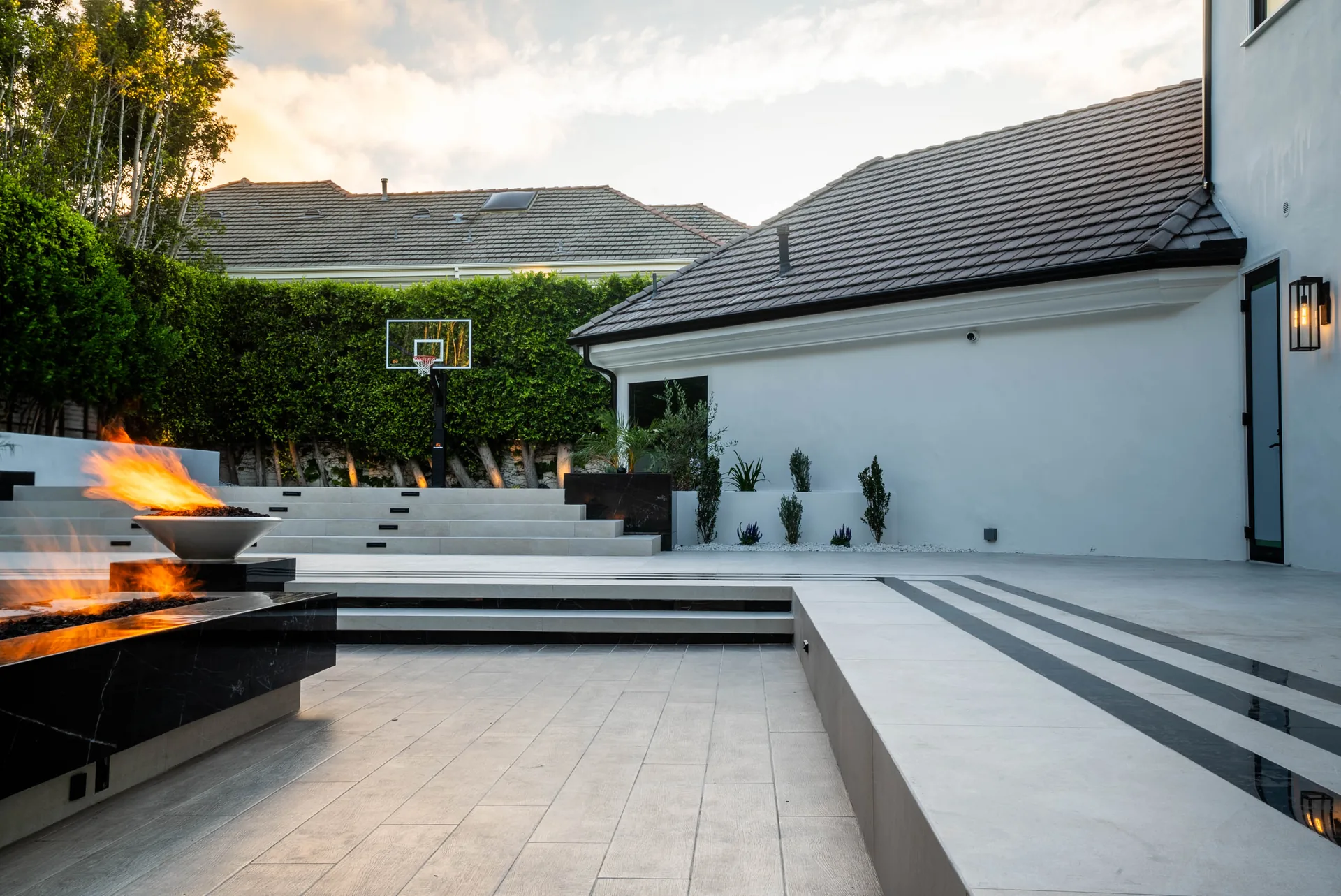 Broad steps running down to the deck, a fire bowl burning at the near corner and striped paving crossing the terrace.