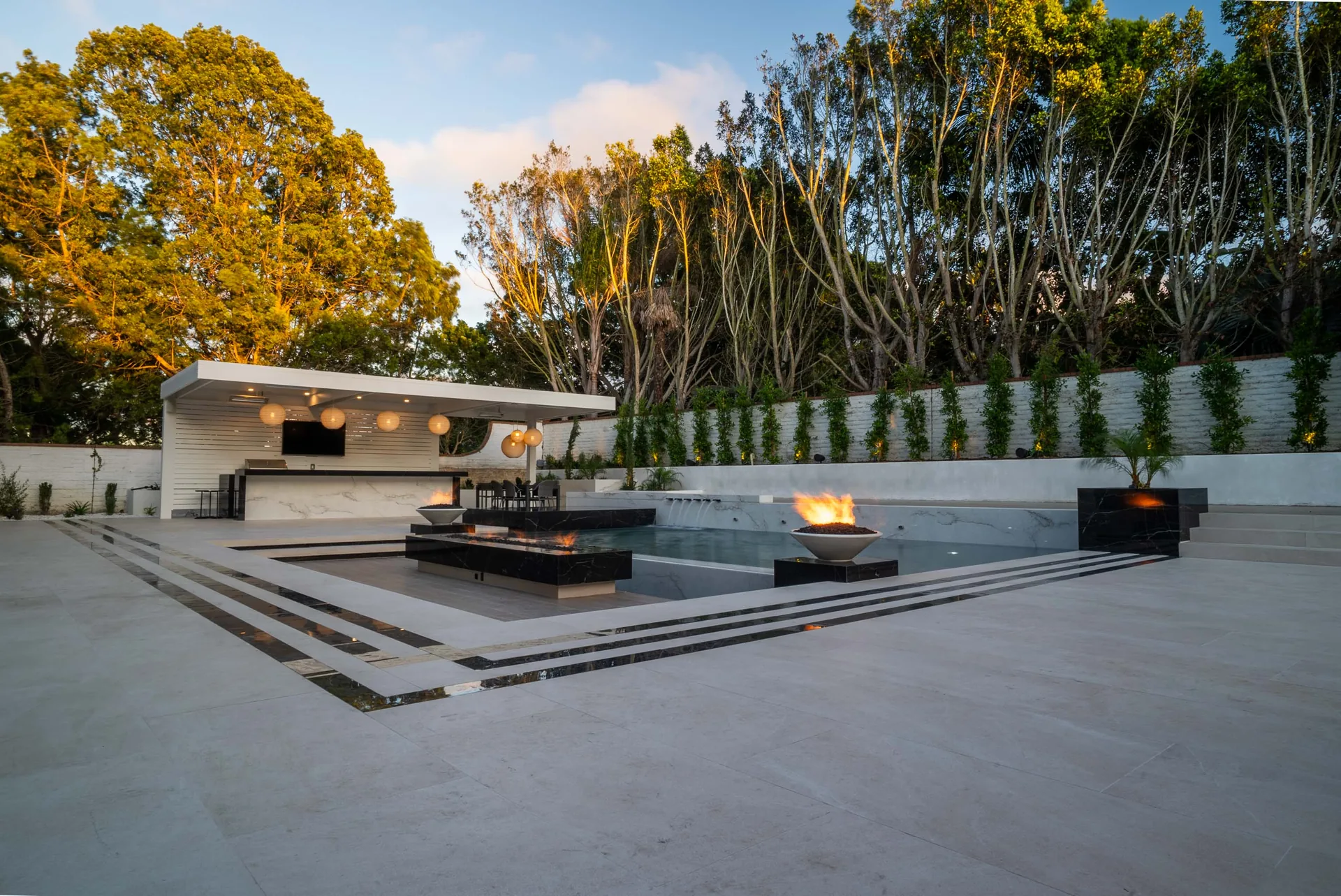 The covered kitchen pavilion seen across the terrace at dusk, fire bowls burning along the pool edge.