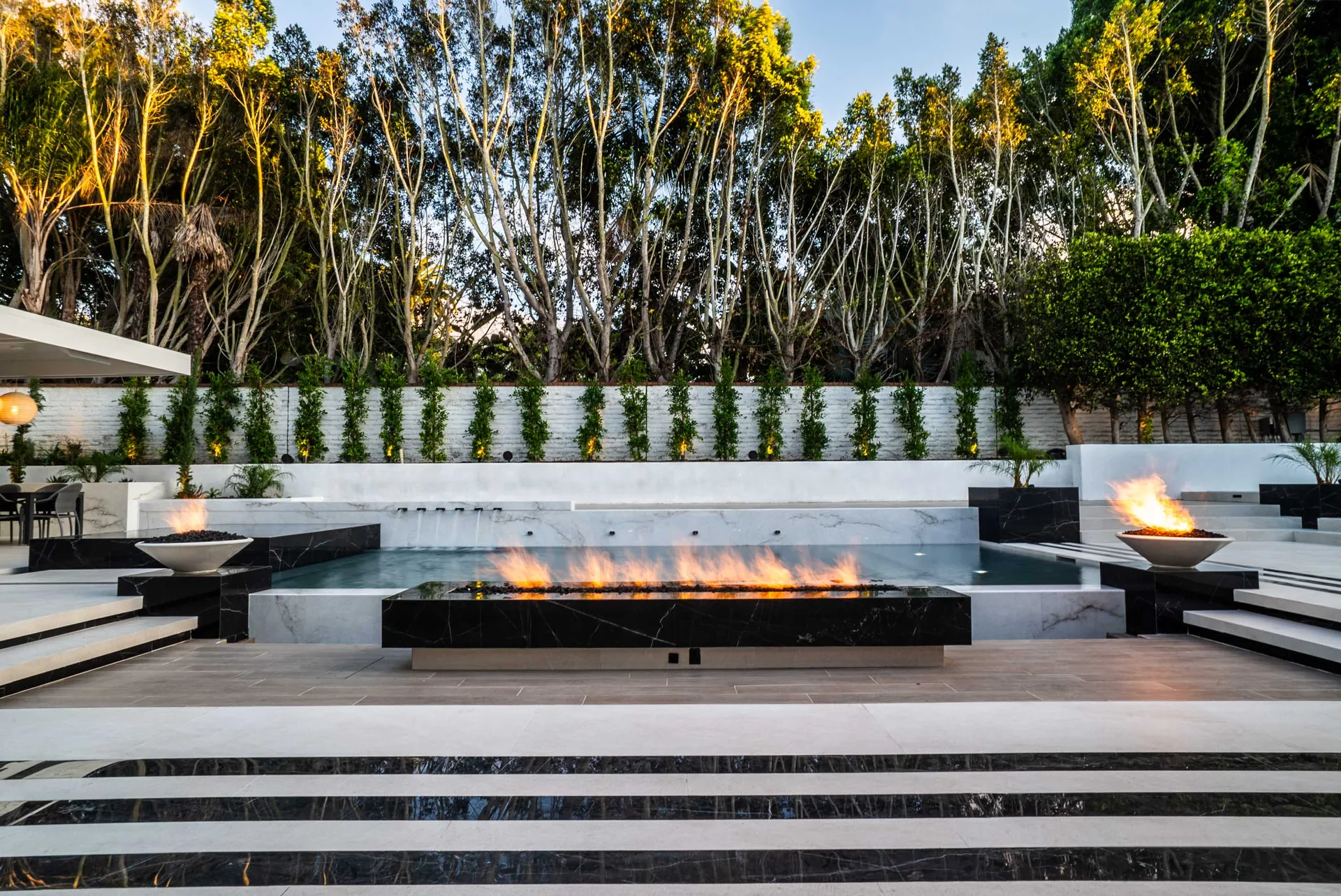 A long linear fire feature burning across the middle of the terrace, flanked by two bowls, with the pool behind.