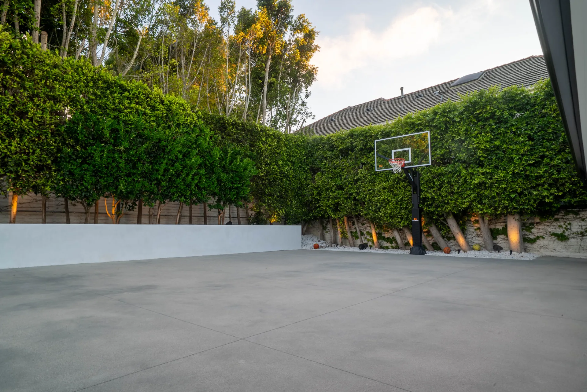 A sport court in pale concrete with a basketball hoop set against a wall of dense green hedge.