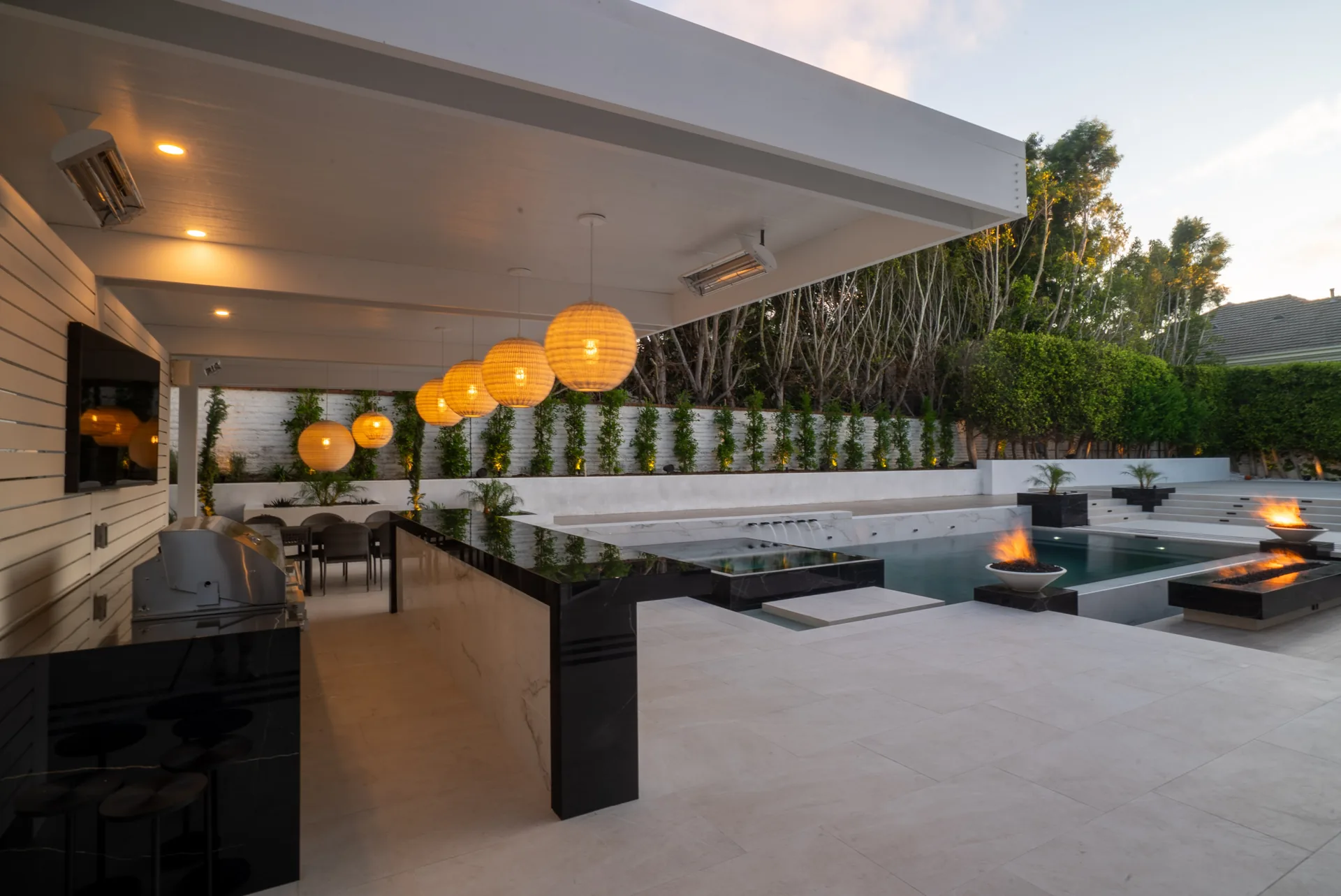 Under the pavilion roof, a marble island, dining table and pendant globes sit with the pool and fire bowls beyond.