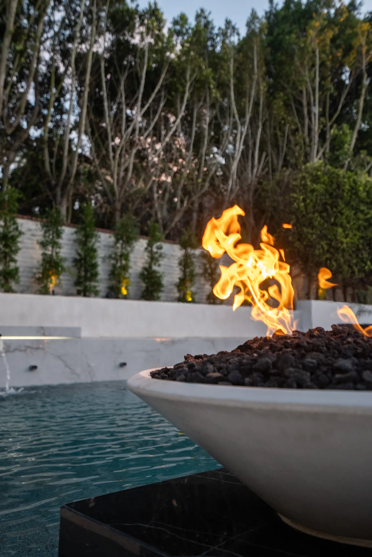Close on a fire bowl burning over black lava rock at the pool's edge, the water dark beneath it.