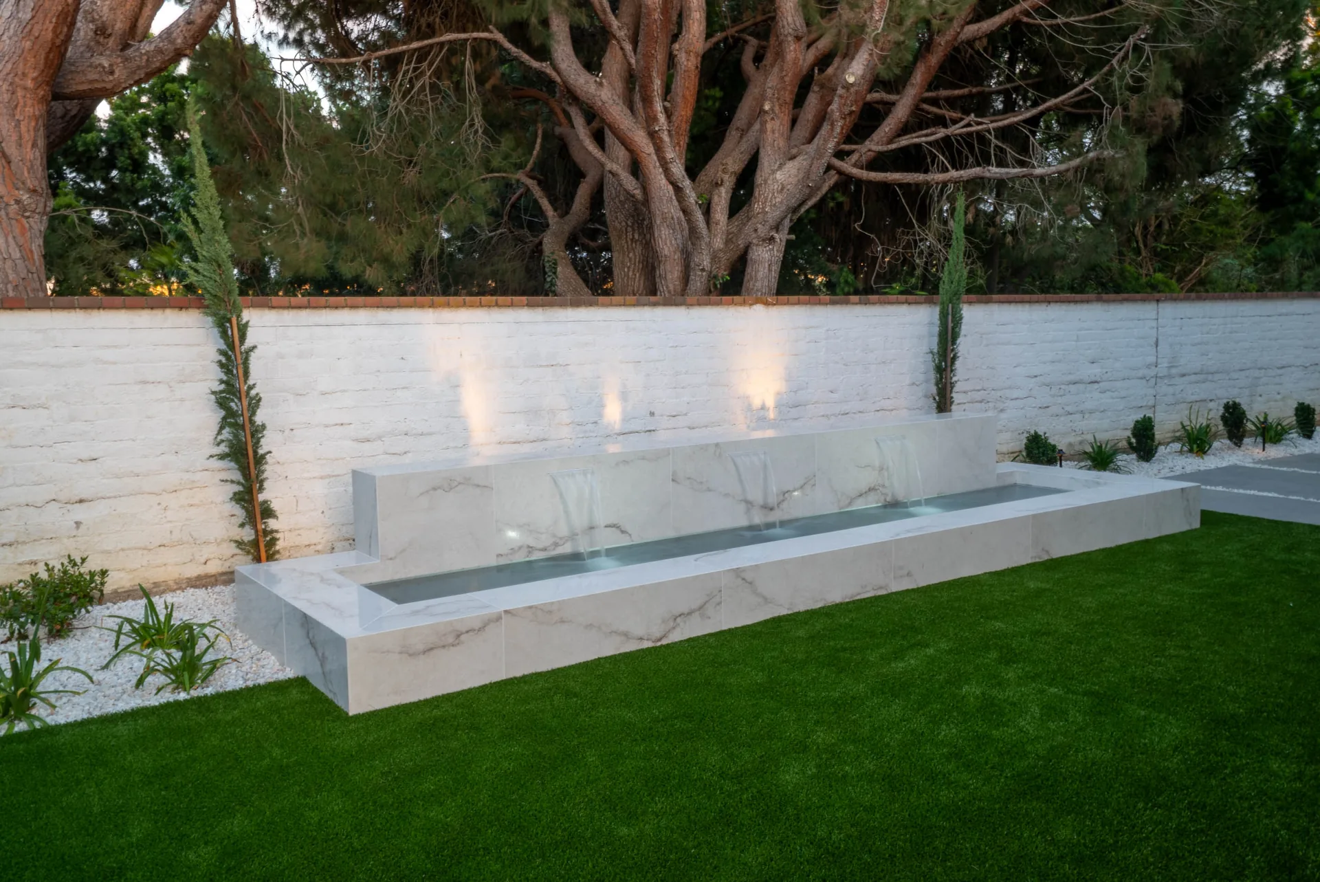 A long white marble water feature running the length of a lawn, uplit against a pale brick wall.